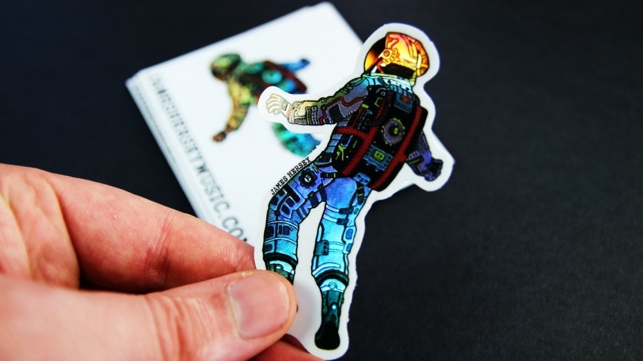 Holographic kiss cut sticker with James Hersey astronaut logo, one peeled and held into the camera