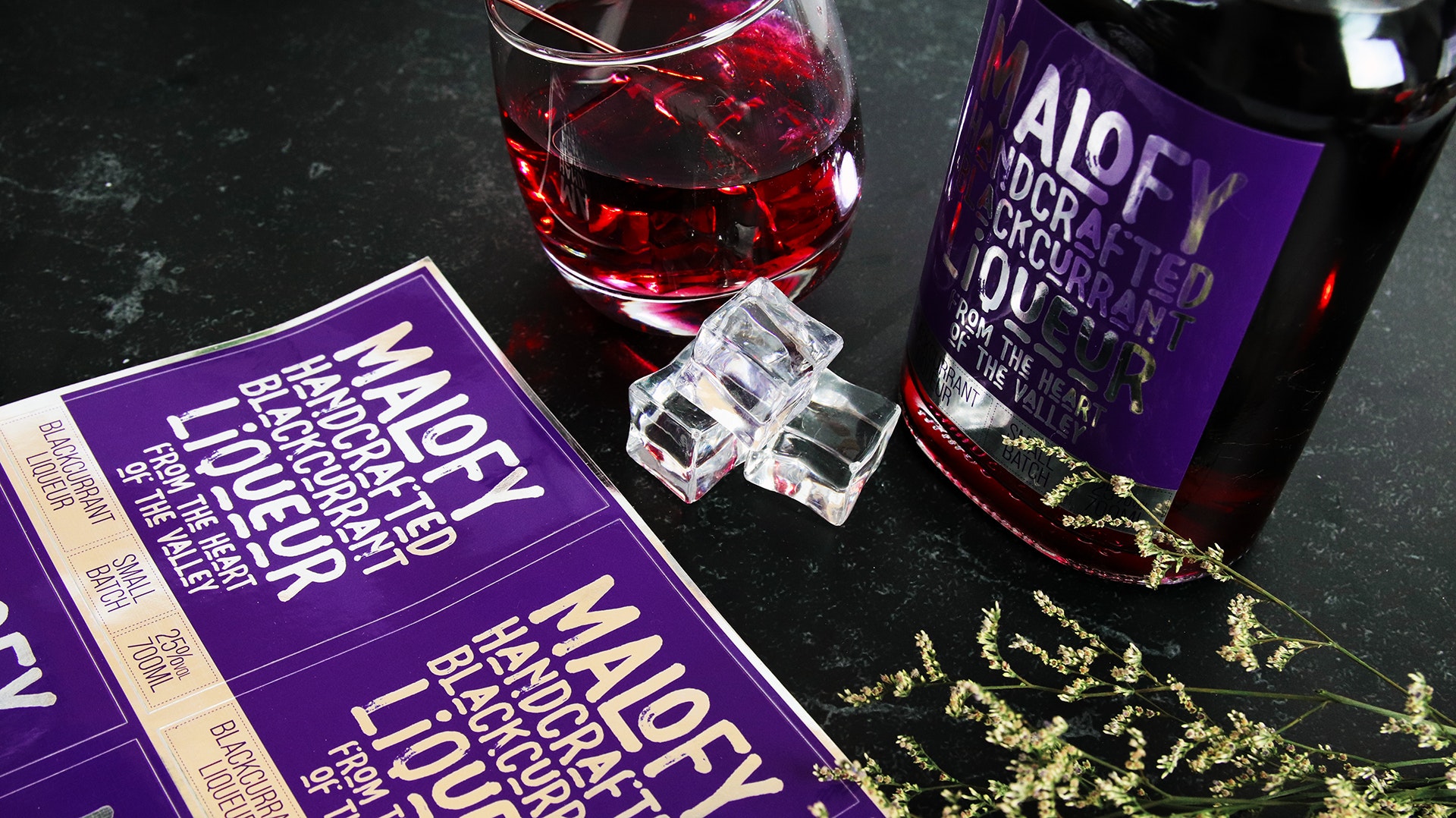 Rectangle mirror silver label applied to a glass bottle containing blackcurrant liqueur with a stack of sticker sheets next to a tilted glass