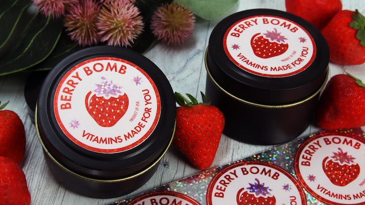 Circle glitter labels applied to black tins and a cardboard box containing a custom made vitamin mix next to glitter sticker sheets