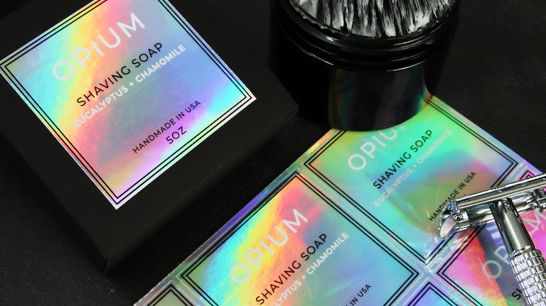 Square holographic sticker applied to black soap box next to shaving tools and sticker sheets