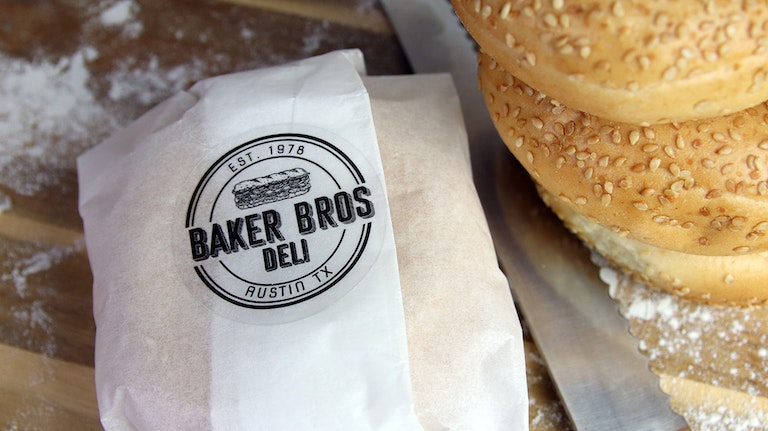 Circle eco-friendly clear sticker with baker bros deli design applied to a white sandwich wrapper next to a stack of bagels