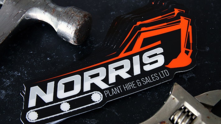 Die cut heavy duty sticker with norris plant hire and sales logo on a black table next to tools
