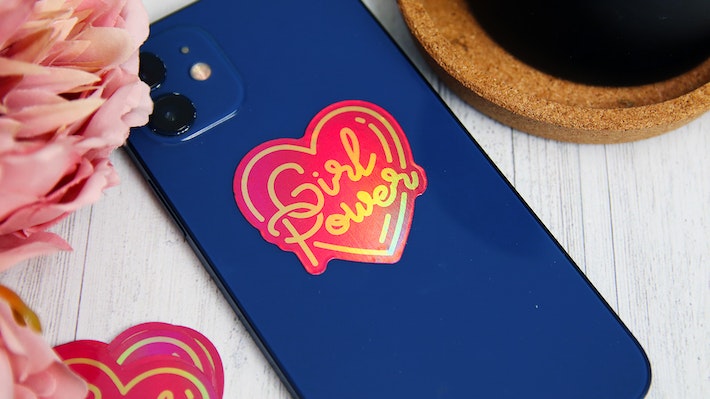 Eco-friendly holographic die cut sticker in the shape of a heart with a girl power logo applied to a blue phone